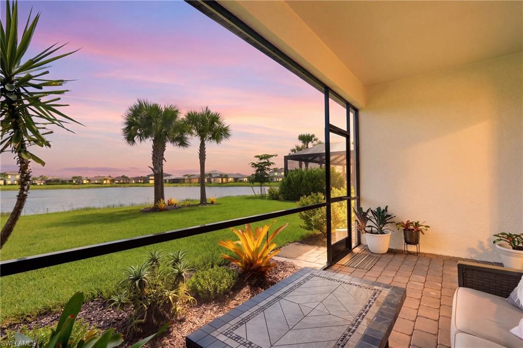 Waterfront patio with sliding glass doors opening to manicured lawn, mature palm trees, tropical landscaping with accent plantings, and serene water views