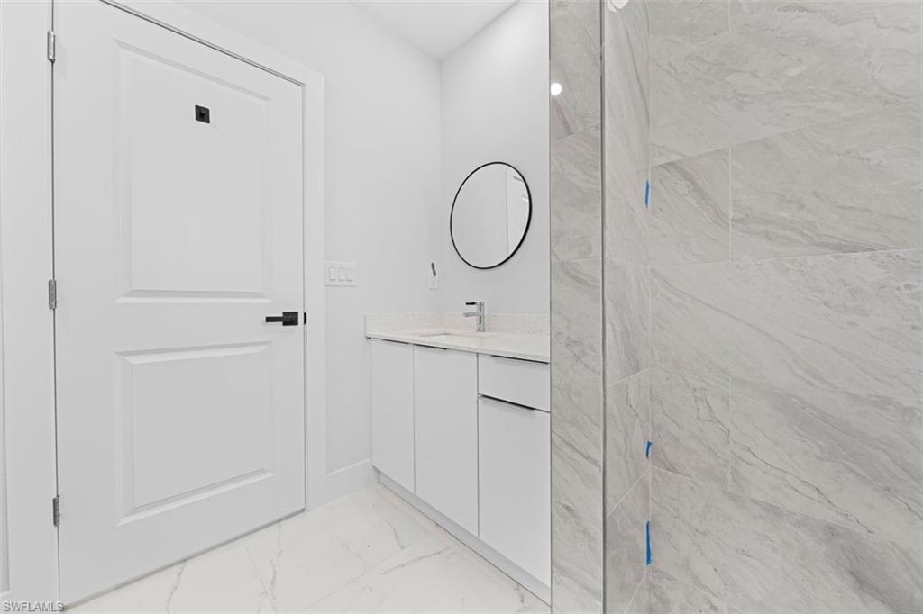 Modern primary bathroom featuring a frameless glass shower enclosure with linear tile detailing, floating vanity with dual drawers and integrated sink, round mirror with metal frame, and light stone-finish wall tile throughout