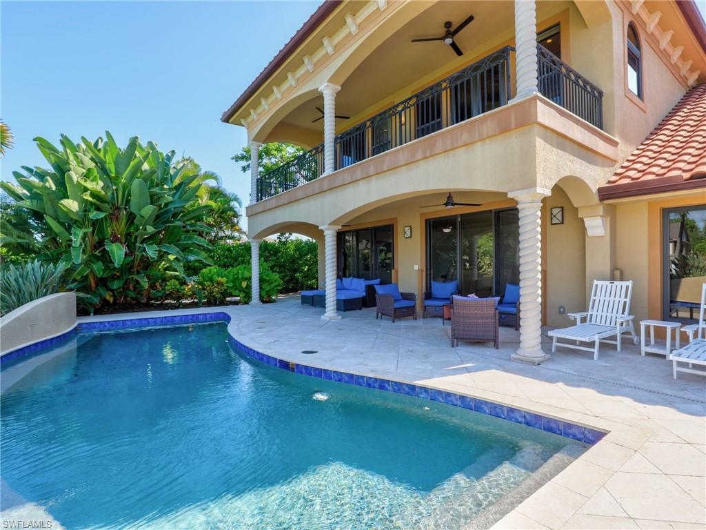 Luxurious resort-style backyard oasis featuring a sparkling saltwater pool with integrated spa, travertine decking, and a two-story Mediterranean-inspired residence with covered loggia, ceiling fans, and French doors opening to the outdoor entertaining space