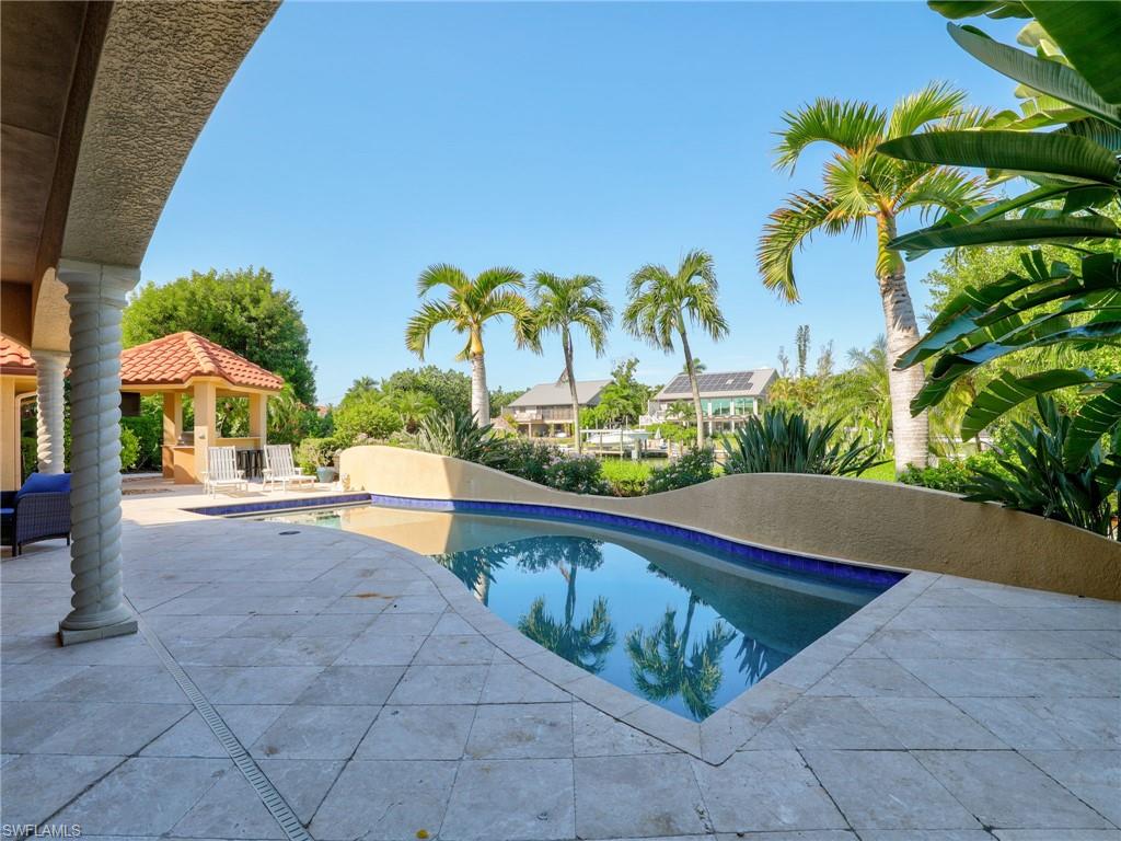 Curved swimming pool with a tile border, extensive paver patio, stone columns, and a built-in gazebo with a tile roof