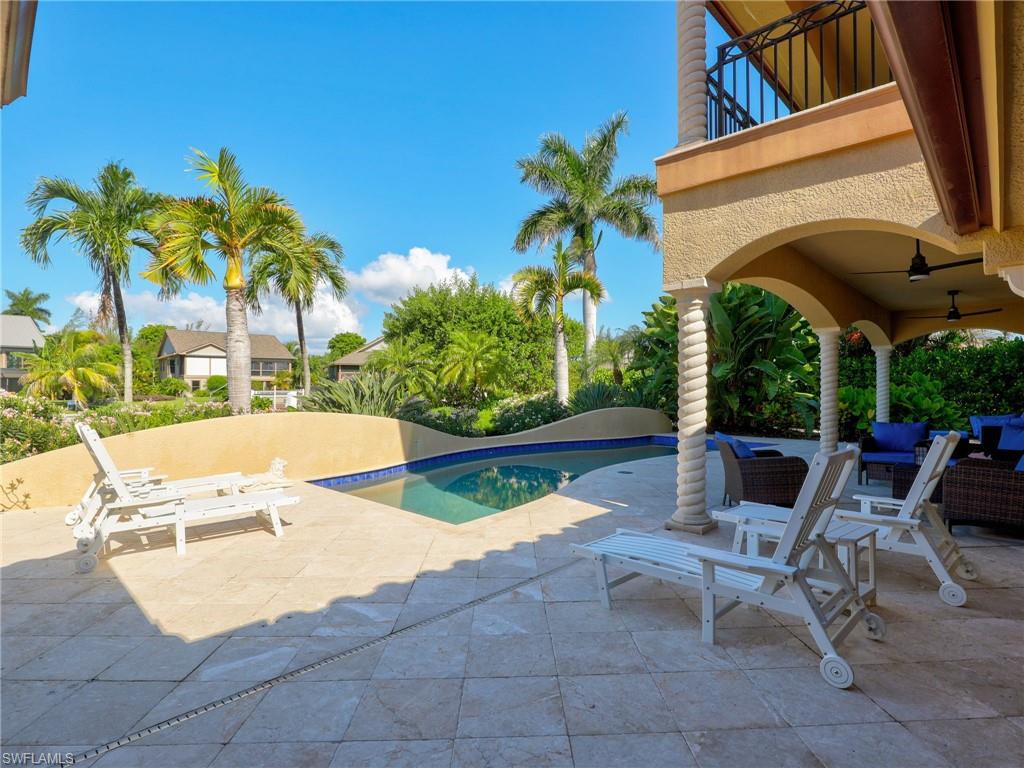 Expansive patio with stone-finish pavers, custom pool with mosaic tile accents, covered outdoor area with archways and ceiling fans, and a second-story balcony with metal railings