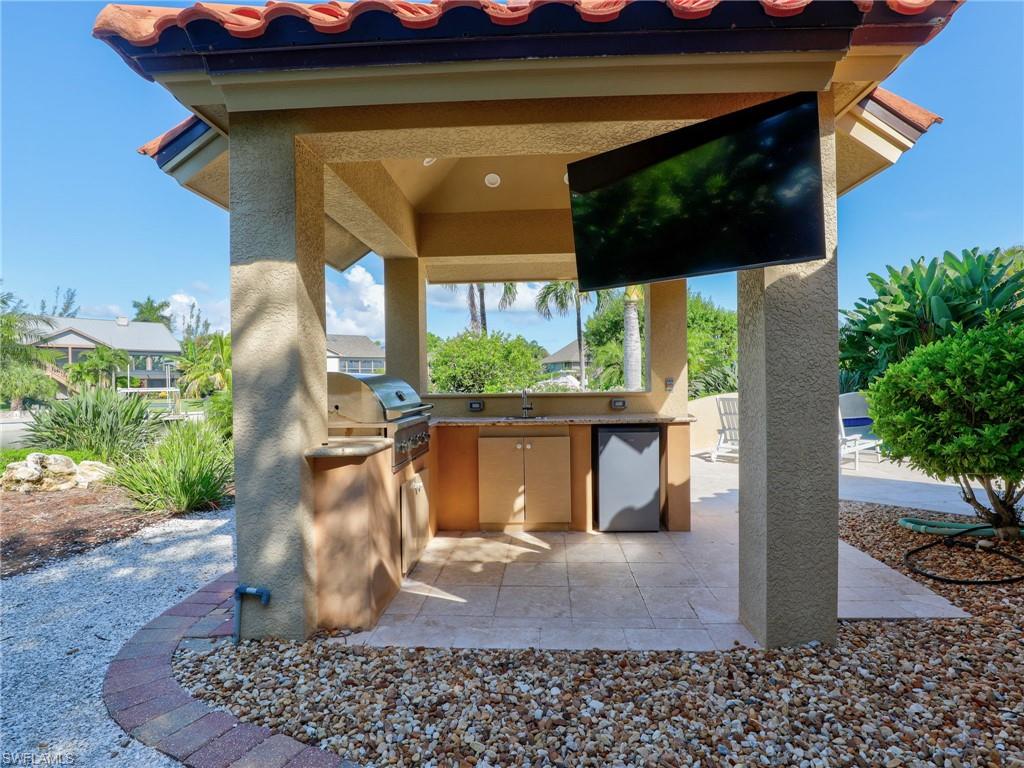 Covered outdoor kitchen featuring a built-in grill, sink, and refrigerator with a television