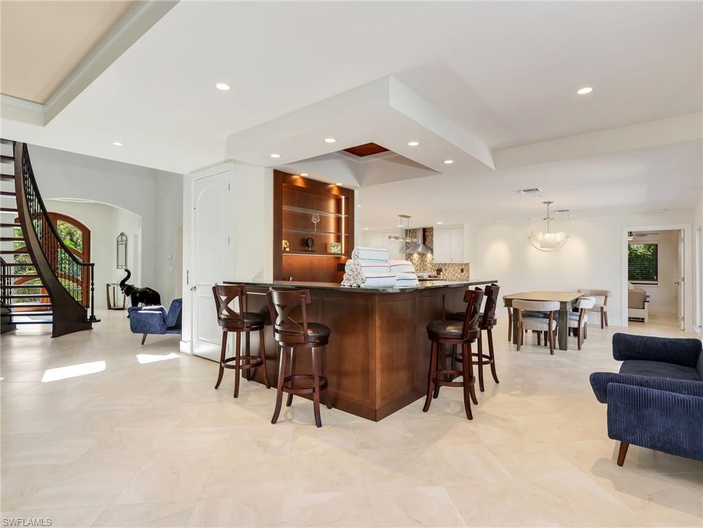 Expansive light-toned tile flooring, grand curved staircase with dark wood and wrought iron accents, built-in wet bar with dark wood cabinetry and integrated shelving, recessed lighting, and a dining area with a contemporary chandelier