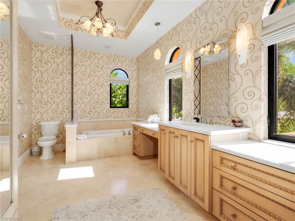 Vaulted ceiling with an ornate chandelier, dual arched windows, built-in soaking tub, extensive vanity with dual sinks, and tile flooring