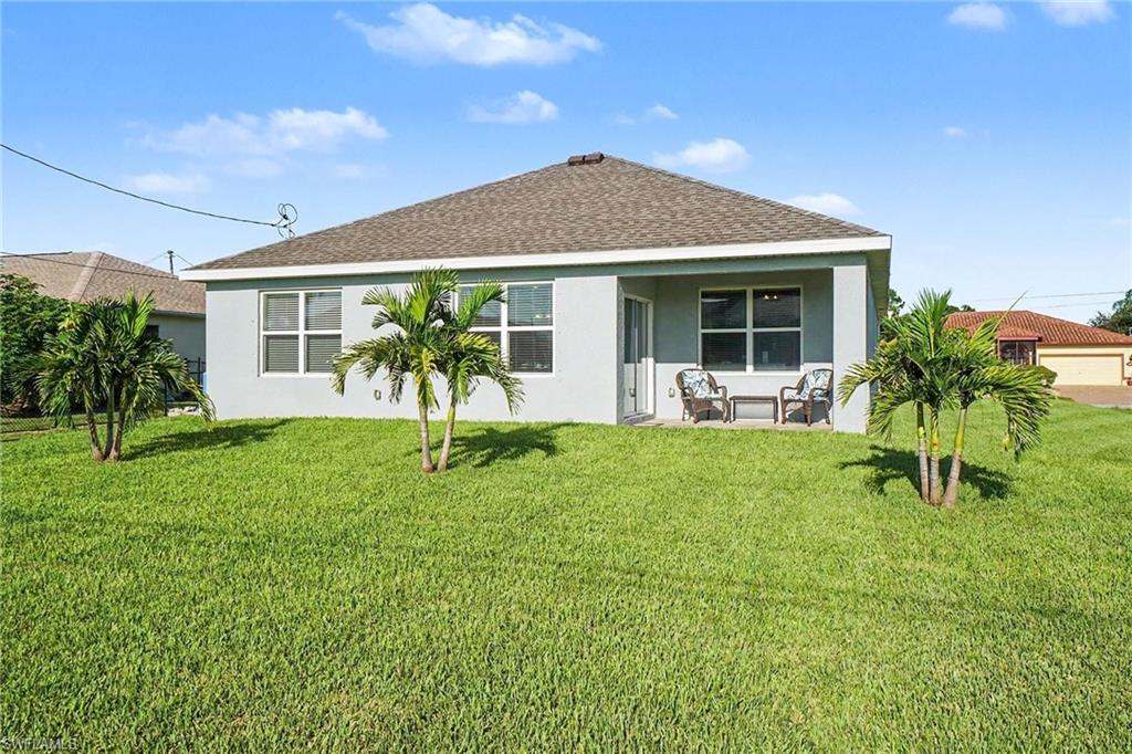 Expansive green lawn, mature palm trees, covered patio, multiple windows, and a shingle roof