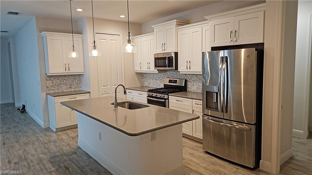 Kitchen featuring an island with an integrated sink, white cabinetry, stainless steel appliances, a tile backsplash, and wood-finish flooring