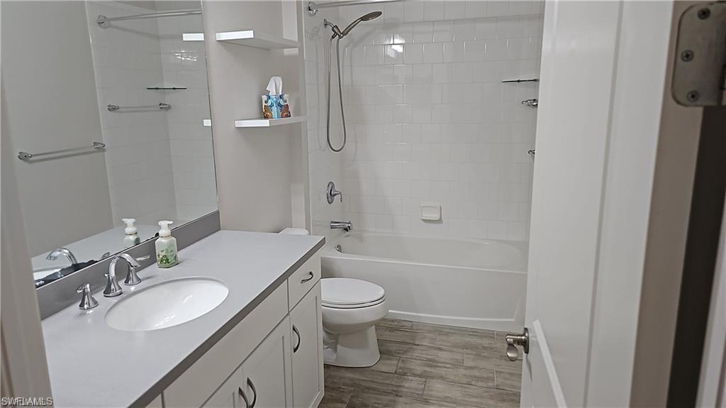 White tiled bath with shower combination, vanity with undermount sink, floating shelves, and wood-finish flooring