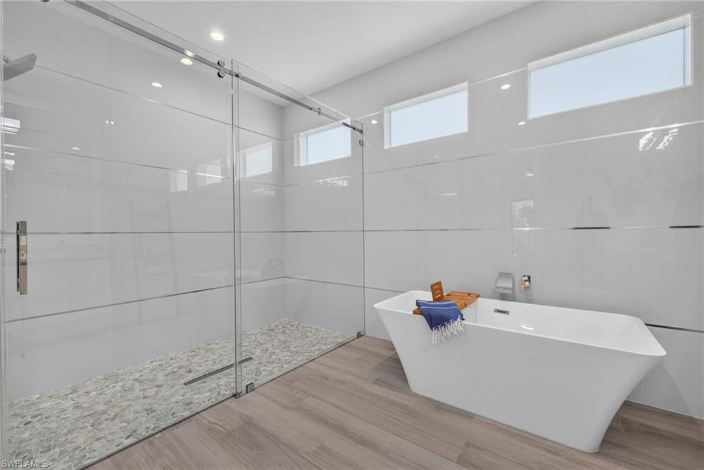 Freestanding soaking tub, glass-enclosed shower with pebble flooring, wood-finish flooring, white tiled walls with horizontal accents, and recessed lighting