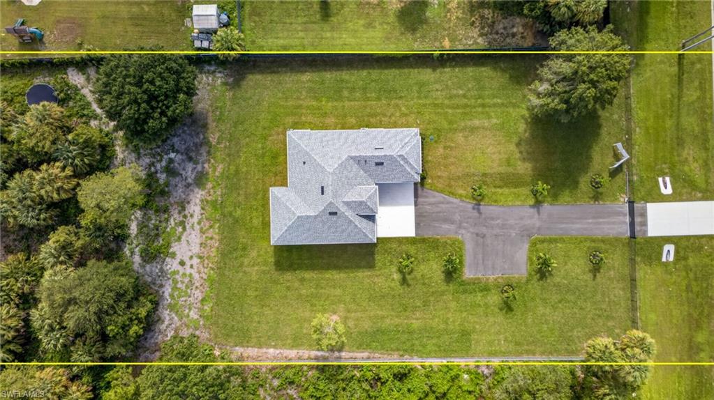 Aerial view of a property featuring a single-story residence, a gray shingle roof, a white concrete driveway, and a paved access road