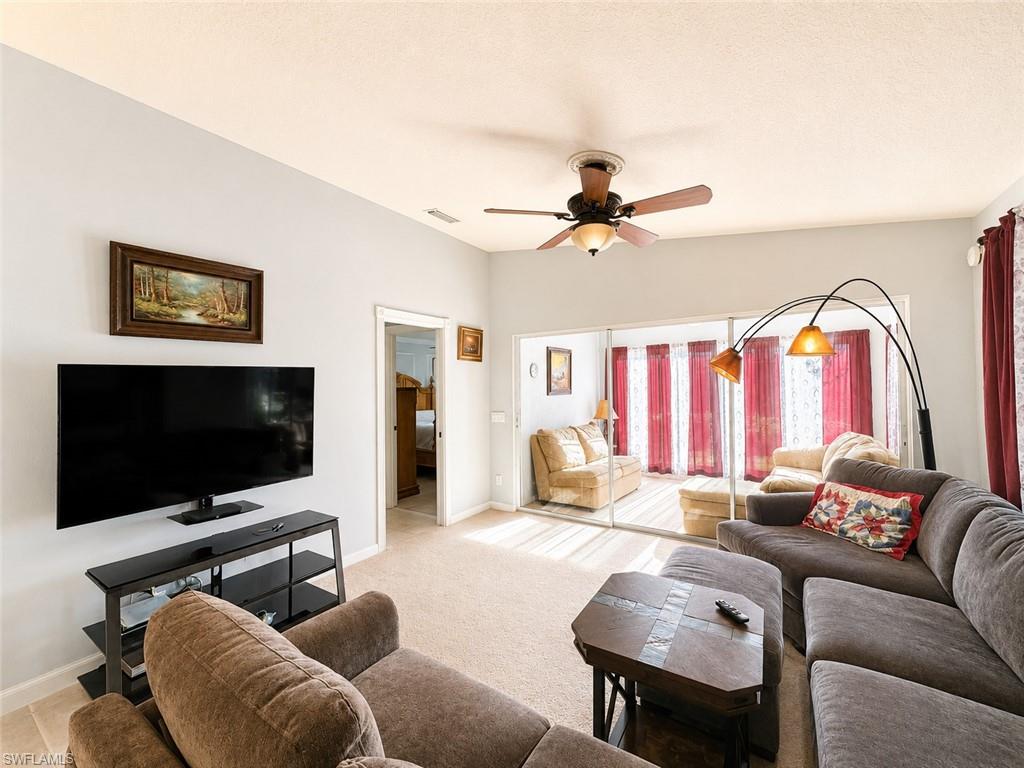 Spacious great room featuring neutral carpet flooring, ceiling fan with integrated lighting, flat-screen television mounted on white wall, and abundant natural light from glass sliding doors