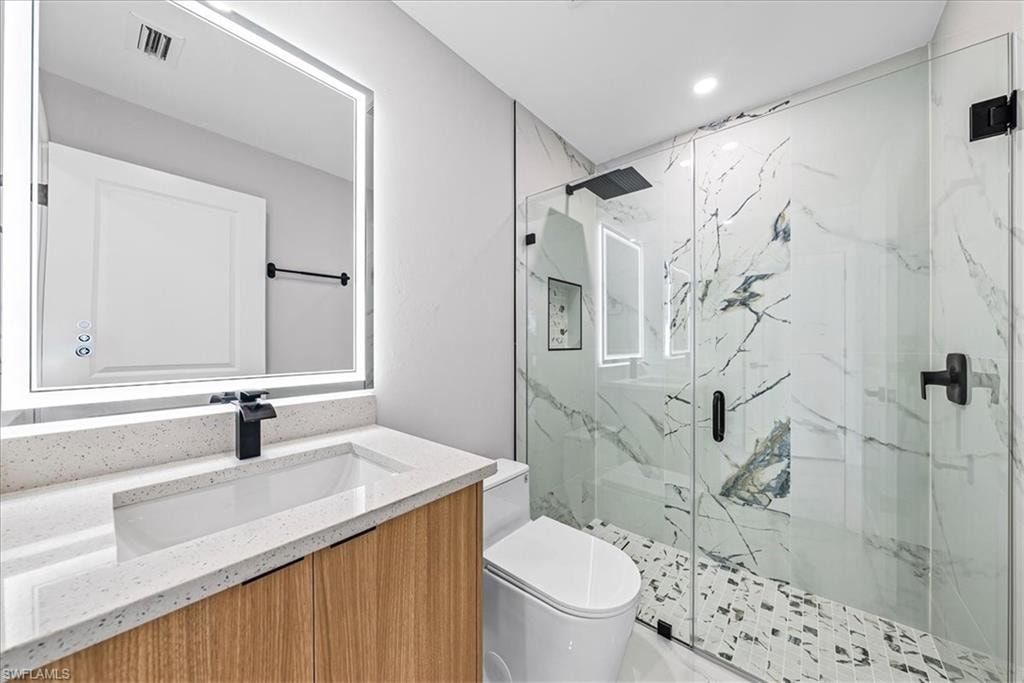 Backlit vanity mirror, wood-finish cabinetry, quartz-finish countertop, glass-enclosed shower, and patterned shower floor tiling