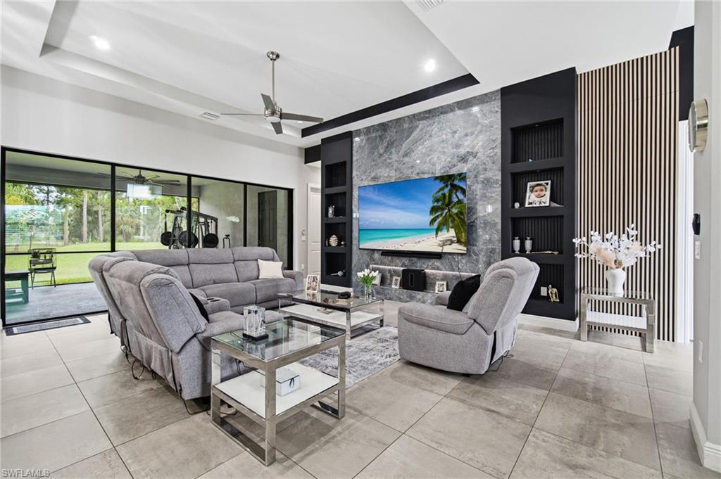 Large format tile flooring, tray ceiling with recessed lighting, ceiling fan, full-wall sliding glass doors, and a decorative accent wall with integrated shelving