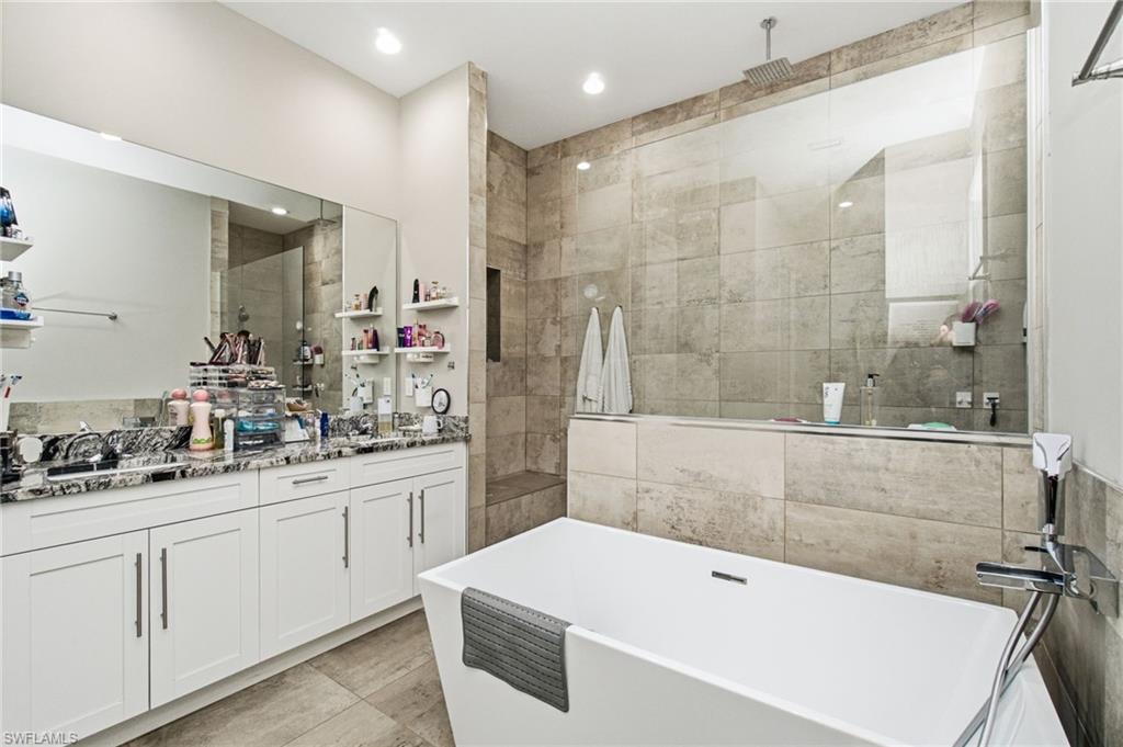 Freestanding soaking tub, walk-in shower with glass enclosure, dual vanity with white cabinetry, and large format tile flooring