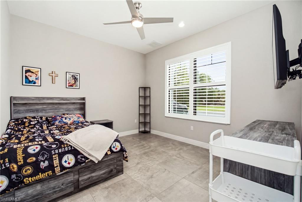 Light gray wall paint with white trim, ceiling fan with integrated lighting, large window with plantation shutters, light gray tile flooring, and a vaulted ceiling