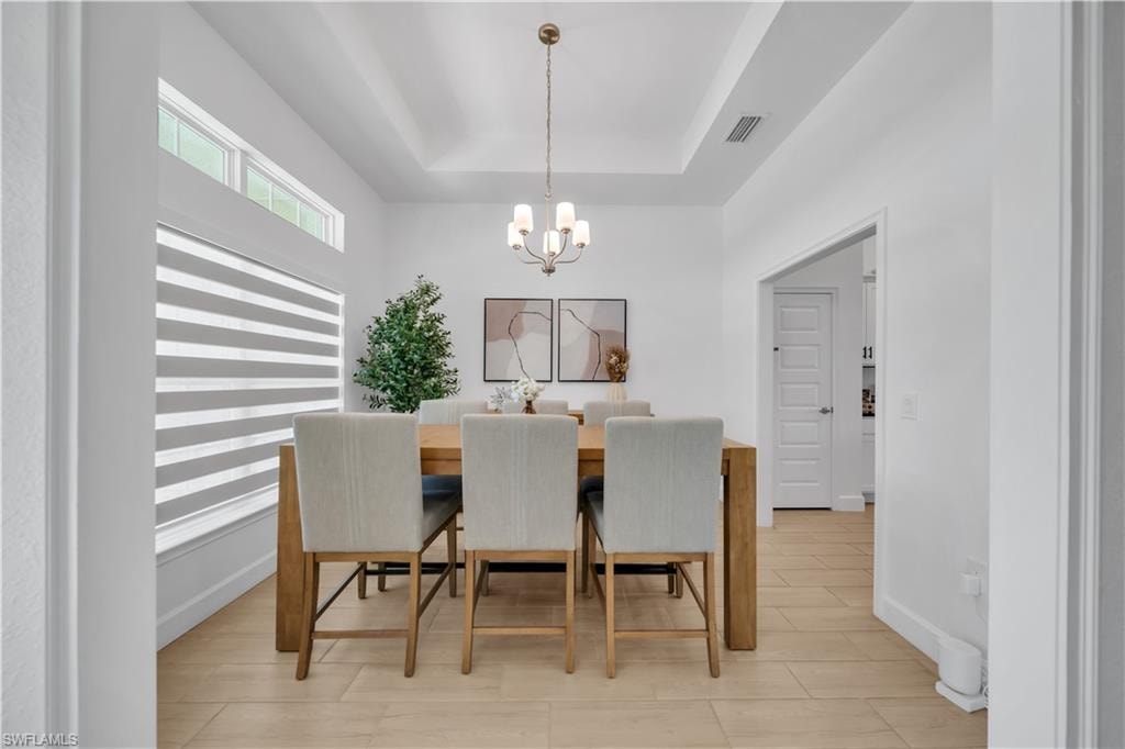 Tray ceiling with a central chandelier, multiple windows with blinds, and light-toned tile flooring