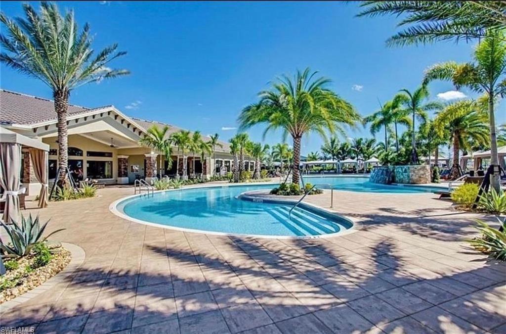 Resort-style pool area with integrated steps, tropical landscaping, paved sundeck, and a prominent building featuring covered entry