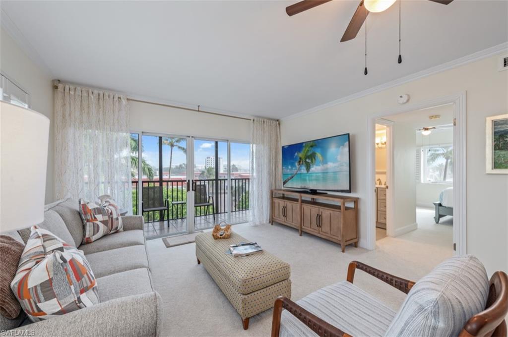 Bright, open living room featuring expansive sliding glass doors with sheer curtains, ceiling fan with pendant lighting, neutral carpet flooring, built-in media console with wood-finish cabinetry, and coastal-inspired artwork