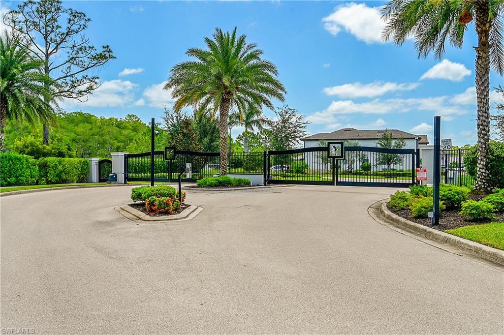 Gated community entrance with asphalt paving, surrounded by mature landscaping and palm trees