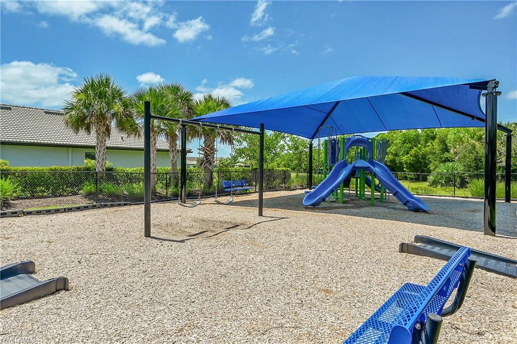Community playground featuring a covered shade structure with blue canopy, modern play equipment including a blue slide tower, ground-level seating with bench-style elements, and mature landscaping with native palms and trees throughout the grounds