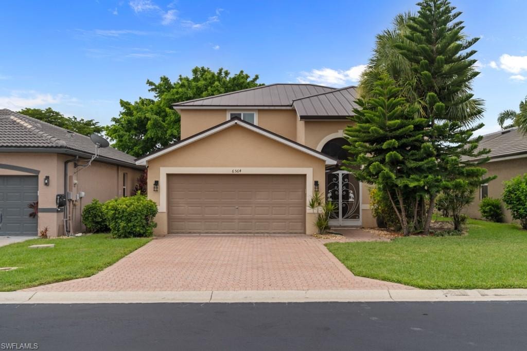Two-story stucco exterior with a metal roof, brick paver driveway, and an arched entry with decorative glass double doors