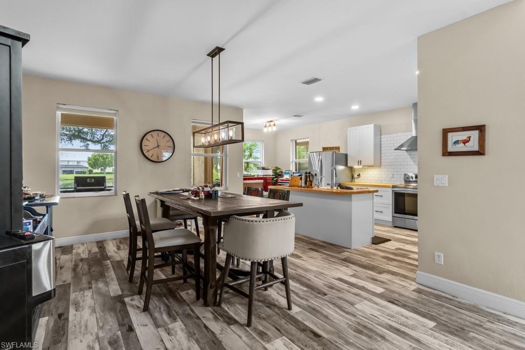 Open-concept dining and kitchen area featuring wood-finish flooring, a dark wood dining table with seating for four, white cabinetry, stainless steel appliances, quartz countertops, and a pendant light fixture suspended from the ceiling