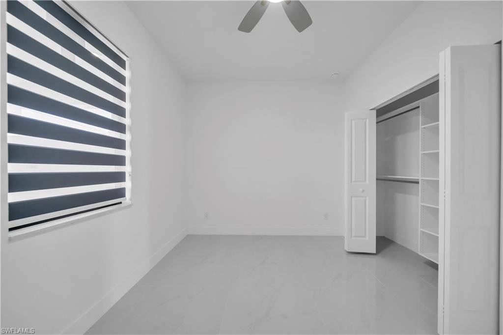 White walls, light-toned flooring, window with layered blinds, built-in closet shelving, and a ceiling fan