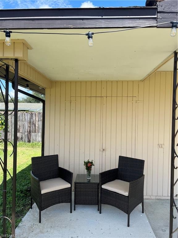Covered concrete patio featuring string lighting, a light-colored siding wall, and a partial decorative metal support column