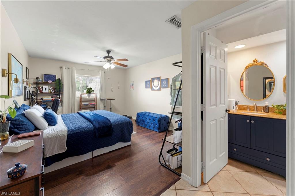 Wood-finish flooring throughout, ceiling fan with light fixture, window with white curtain panels, solid panel interior door, and a bathroom featuring a vanity with a wood-finish countertop and gold-framed mirror