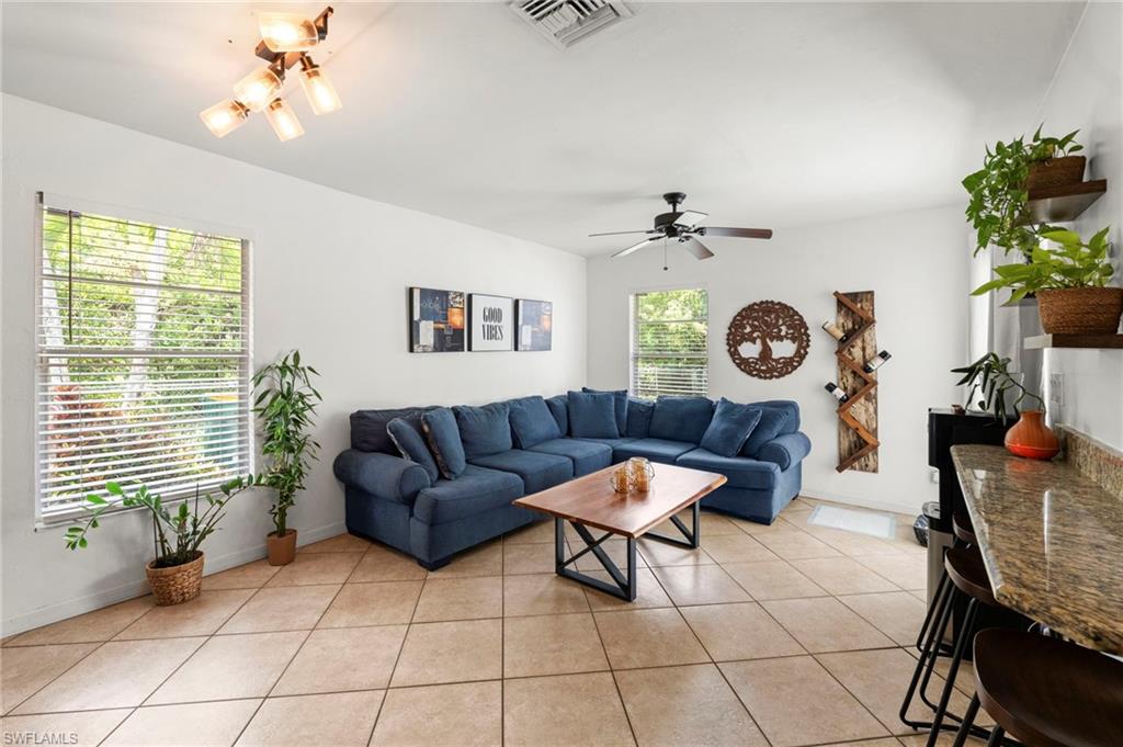 Spacious living area featuring neutral tile flooring, a ceiling fan, multiple windows, and a breakfast bar with a granite-style countertop