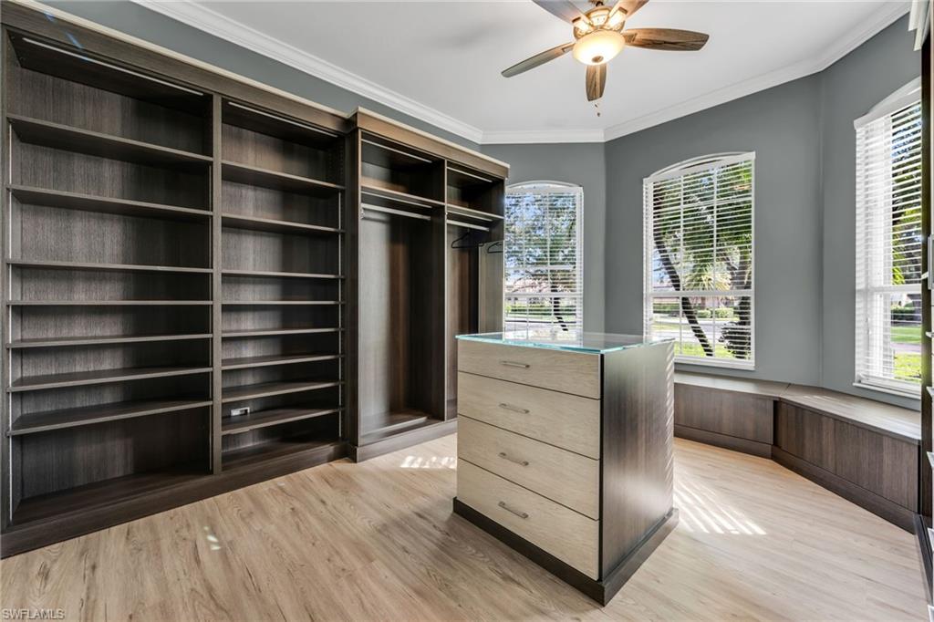 Built-in shelving and cabinetry with hanging rods, wood-finish flooring, central storage island with glass top, arched windows with blinds, and a ceiling fan with integrated lighting