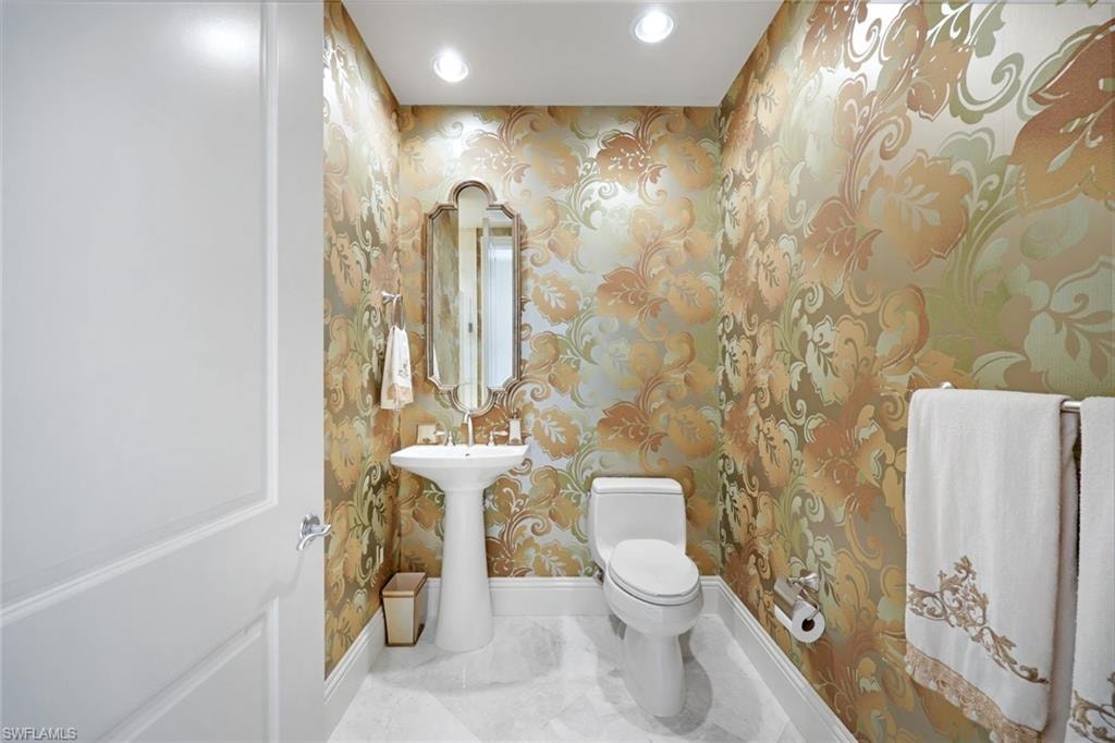 Pedestal sink with polished chrome fixtures, ornate wall-mounted mirror, decorative patterned wallpaper, recessed ceiling lighting, and white tile flooring