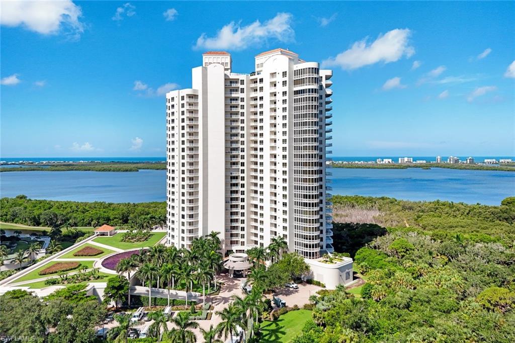 Waterfront high-rise residential tower featuring contemporary white exterior, multiple stories with extensive fenestration, manicured landscaping with mature palm trees, and direct water and natural preserve vistas from an elevated position