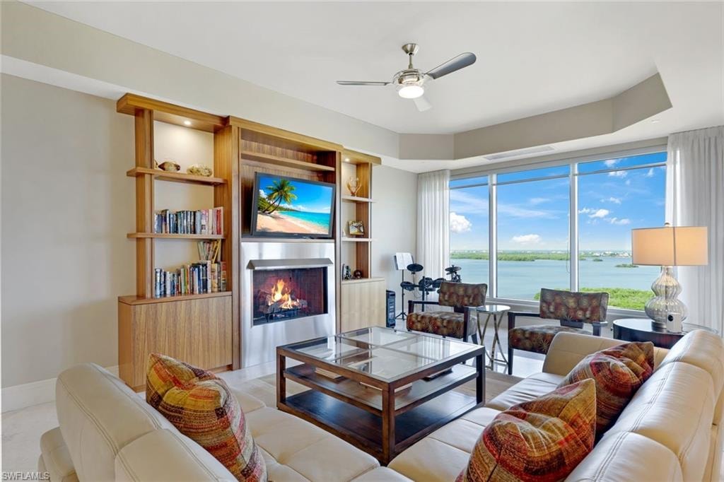 Sophisticated living room featuring floor-to-ceiling windows overlooking expansive waterfront scenery, custom wood cabinetry with integrated fireplace and entertainment shelving, ceiling fan, neutral upholstered seating, dark wood coffee table, and table lamps with warm lighting