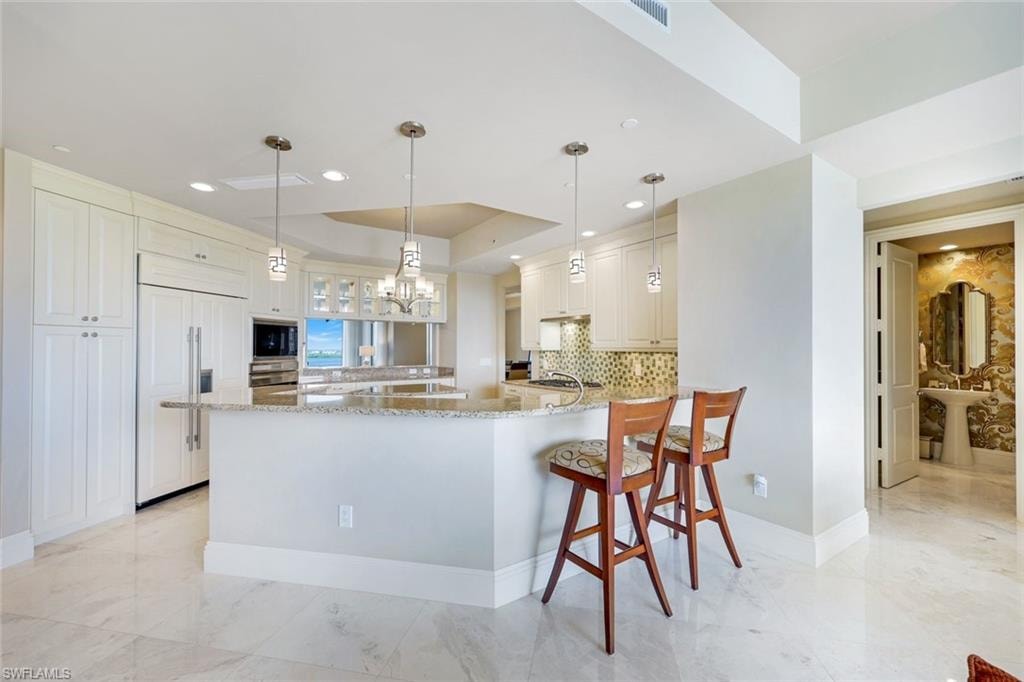 Gourmet kitchen featuring a large waterfall-edge island with seating for two, stainless steel appliances, white cabinetry, pendant lighting, and polished stone flooring throughout the open living area