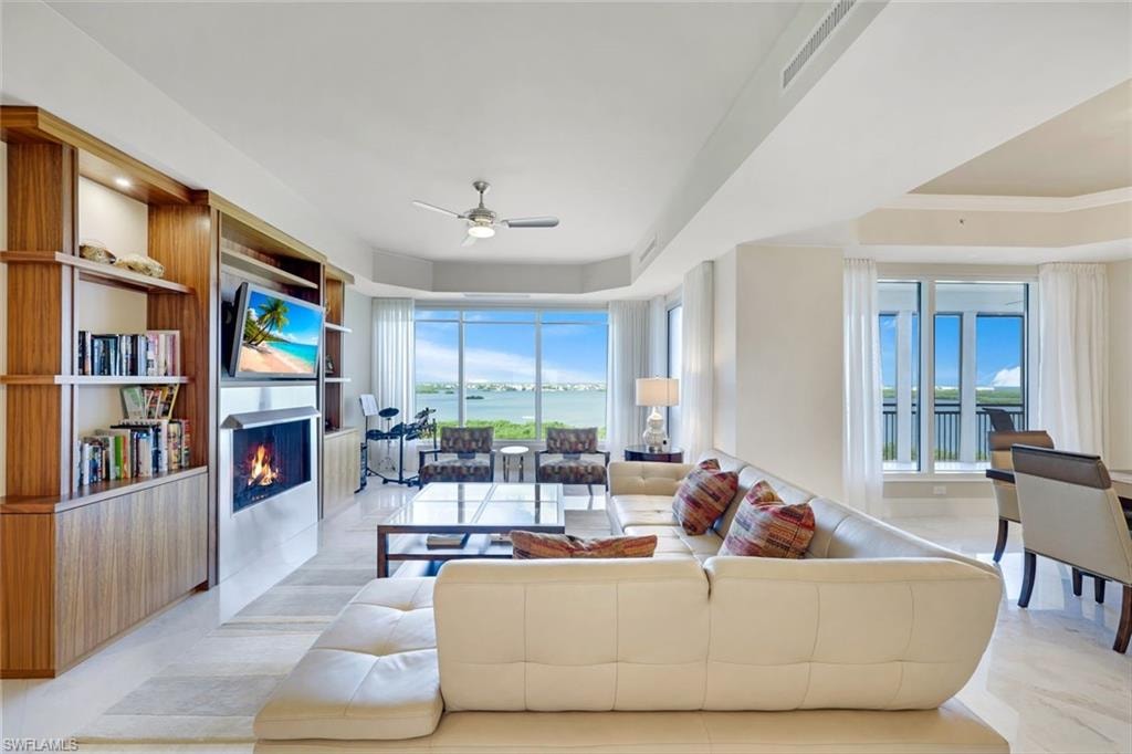 Sophisticated open-concept living space showcasing floor-to-ceiling windows framing expansive water and landscape vistas, curved cream-upholstered seating, wood-finish cabinetry with integrated fireplace and media storage, light-toned flooring throughout, and ceiling fan for climate control