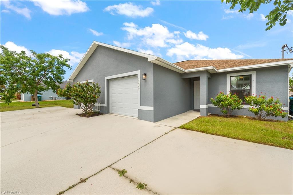 Stucco exterior with a shingled roof, attached garage, concrete driveway, and established landscaping