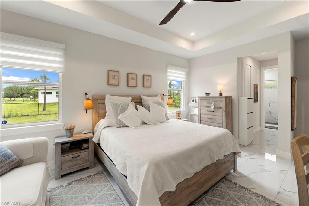Spacious primary bedroom featuring a wood-frame bed, dual large windows with plantation shutters flooding the space with natural light, tile flooring throughout, ceiling fan, built-in nightstands with storage, and an ensuite bathroom visible through the adjacent doorway