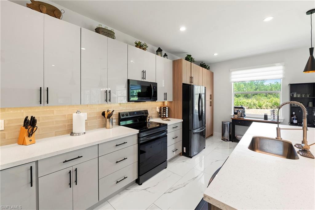 Modern kitchen featuring white cabinetry with black hardware, quartz countertops, stainless steel appliances including a black range and refrigerator, a subway tile backsplash, an oversized island with a copper sink, and abundant natural light from large windows overlooking the landscaped grounds