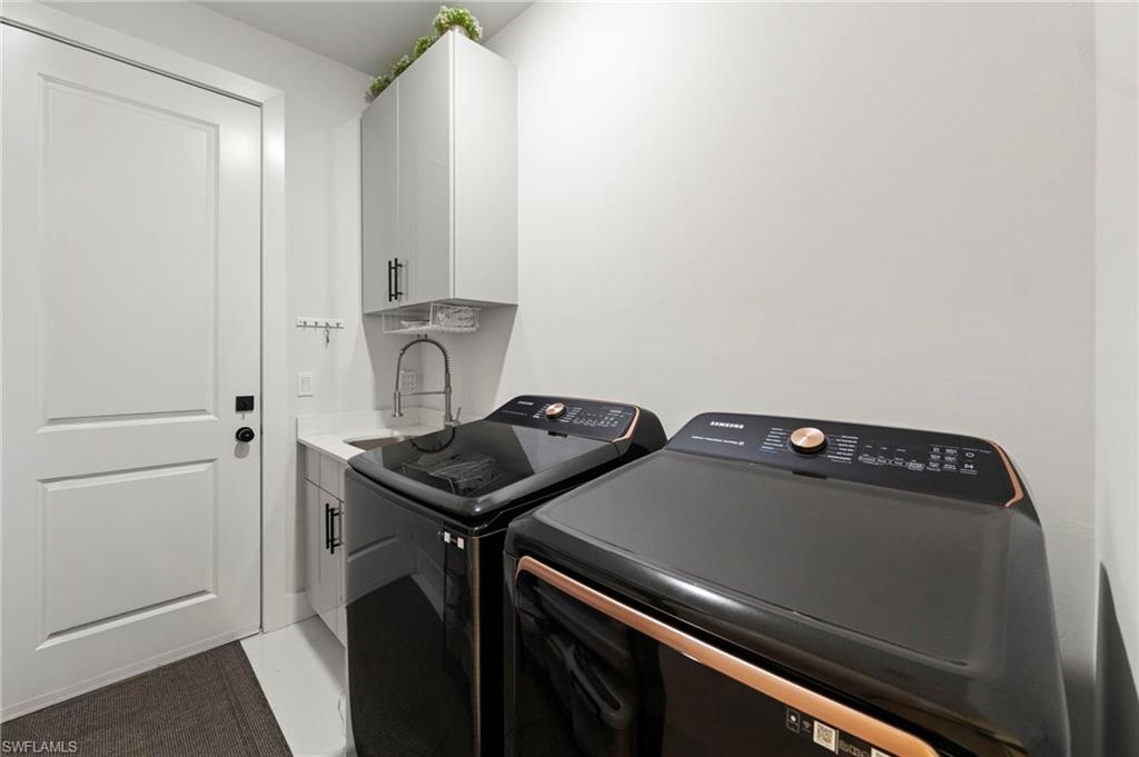 Modern laundry room featuring side-by-side black front-load washer and dryer with copper accents, white cabinetry with integrated sink and gooseneck faucet, white subway tile backsplash, and direct entry from the main living space