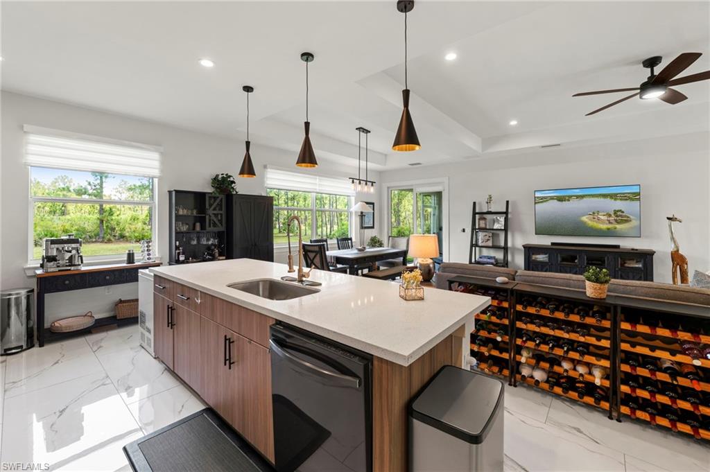 Open-concept kitchen and living area featuring a large wood-finish island with white quartz countertop and integrated cooktop, stainless steel appliances, pendant lighting with bronze fixtures, ceiling fan, recessed lighting, and expansive windows overlooking landscaped grounds