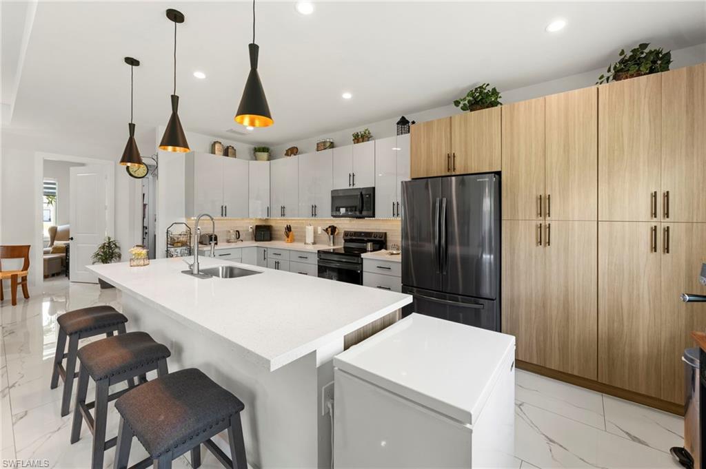 Contemporary kitchen featuring a large waterfall-edge quartz island with seating for multiple guests, stainless steel appliances including a side-by-side refrigerator, white cabinetry with natural wood-finish upper storage, pendant lighting with brass accents, and polished porcelain flooring throughout the open layout