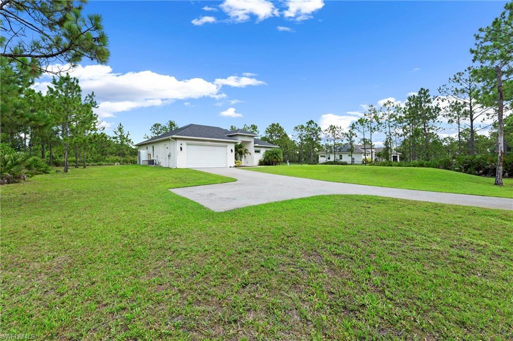 Expansive grounds featuring manicured lawn with mature tree-lined perimeter, concrete driveway approach, and modern single-story residence with neutral exterior finishes set on substantial acreage under clear skies