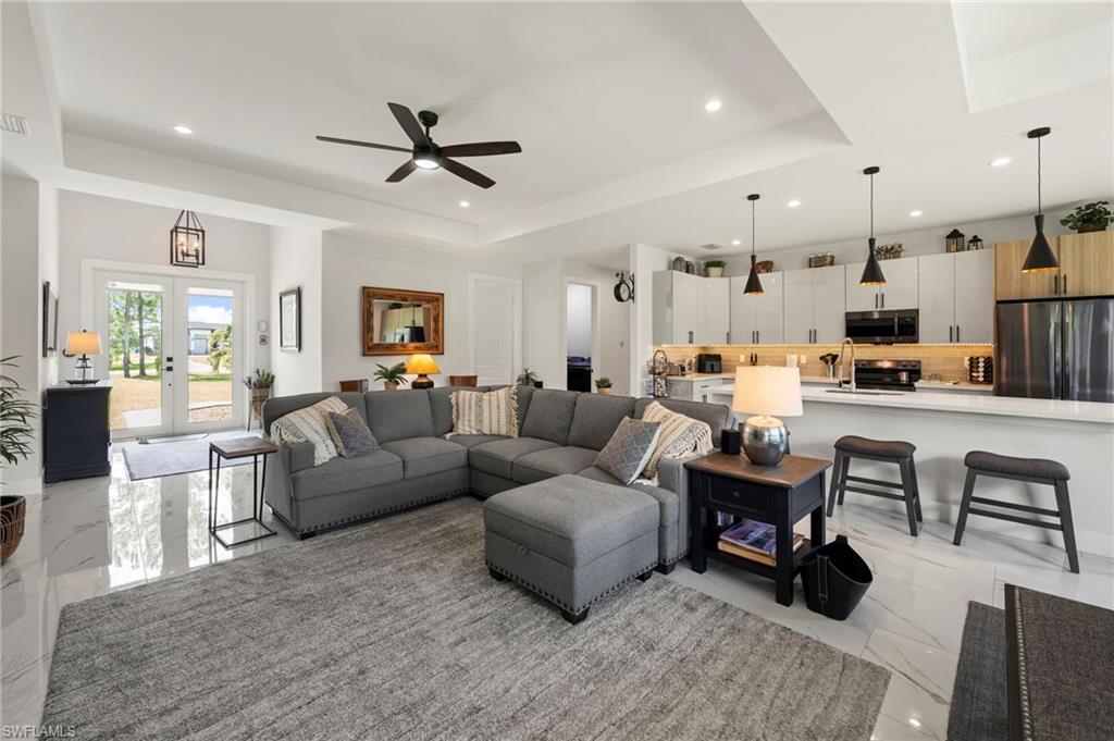 Open-concept living and kitchen featuring a gray upholstered sectional with ottoman, stainless steel appliances, white cabinetry with wood-finish accents, pendant lighting over the kitchen island, ceiling fan, and polished light-toned flooring throughout the space