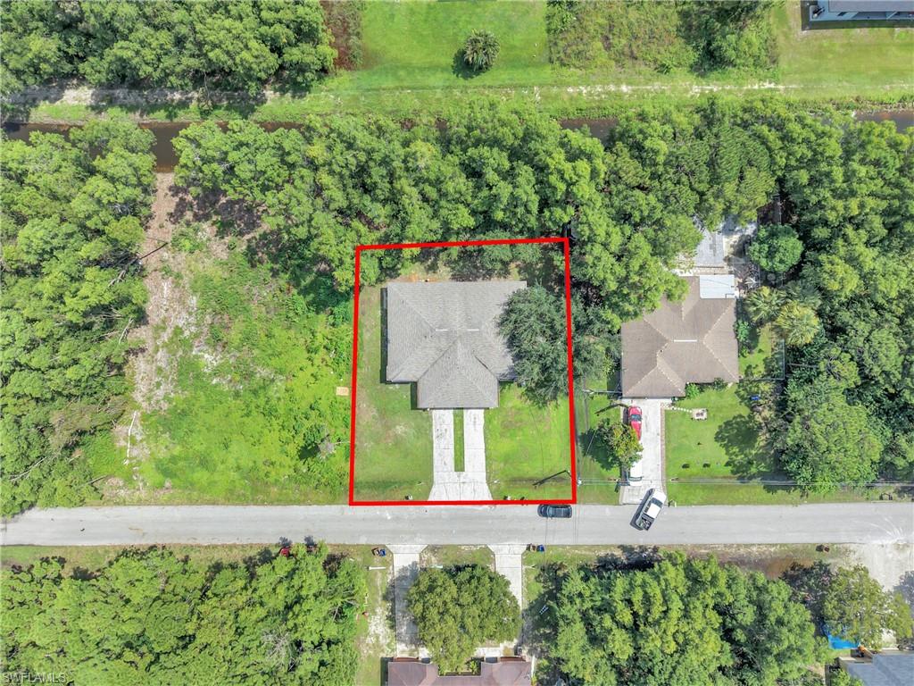 Aerial property overview featuring a single-story residence with a gabled roof, an extended concrete driveway, and surrounding mature trees