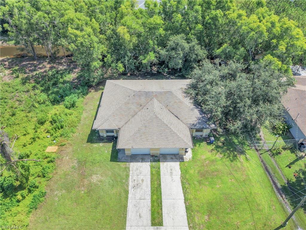 Residential property with dual asphalt shingle roofs, surrounded by mature trees, featuring a concrete driveway, and a large grassy yard