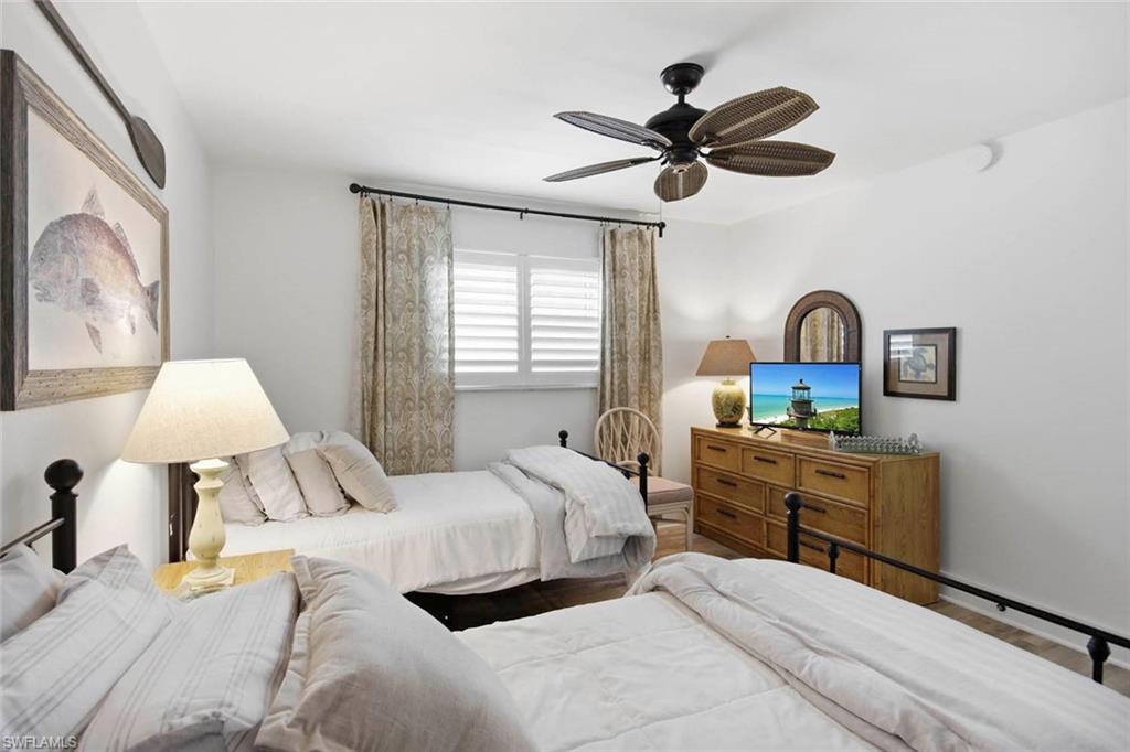 Bright secondary bedroom with dual twin beds, wood-finish dresser with integrated media station, ceiling fan with wood-grain blades, and natural light from shuttered windows dressed with neutral linen panels