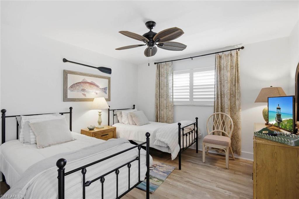 Bright guest bedroom featuring two wrought-iron bed frames with black finials, wood-finish flooring, ceiling fan with dark finish, and large windows with plantation shutters and light-filtering curtains