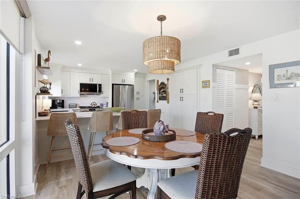 Open-concept dining and kitchen area featuring a round white pedestal dining table, woven rattan chairs, wood-finish flooring, stainless steel appliances, white cabinetry with hardware accents, and a woven pendant light fixture suspended from the ceiling