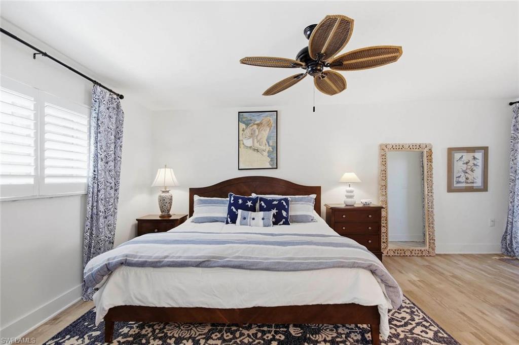 Primary bedroom featuring a wood-finish ceiling fan with integrated lighting, wood-framed bed with upholstered headboard, wood-finish flooring, dual nightstands with table lamps, decorative mirror with woven frame, and windows with blue patterned curtains and plantation shutters