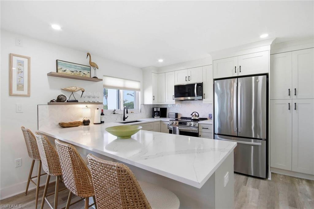 Modern kitchen featuring white cabinetry with hardware accents, a substantial quartz-topped island with waterfall edge, stainless steel appliances including a side-by-side refrigerator and gas cooktop, and light wood-finish flooring throughout